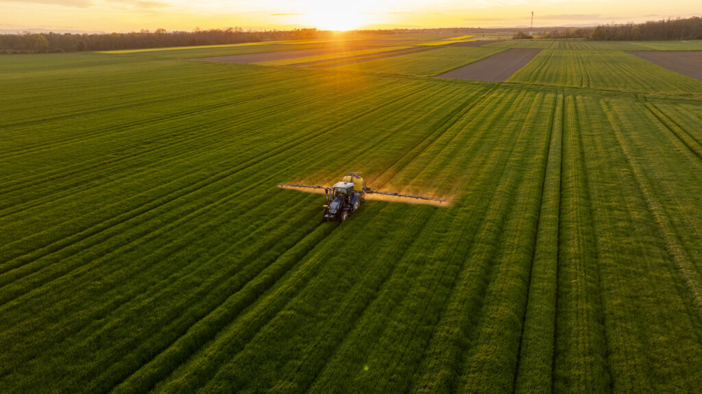 A tractor is fertilizing a vast green agricultural field at sunset, creating a serene rural atmosphere. The golden light adds warmth and depth to the lush landscape.